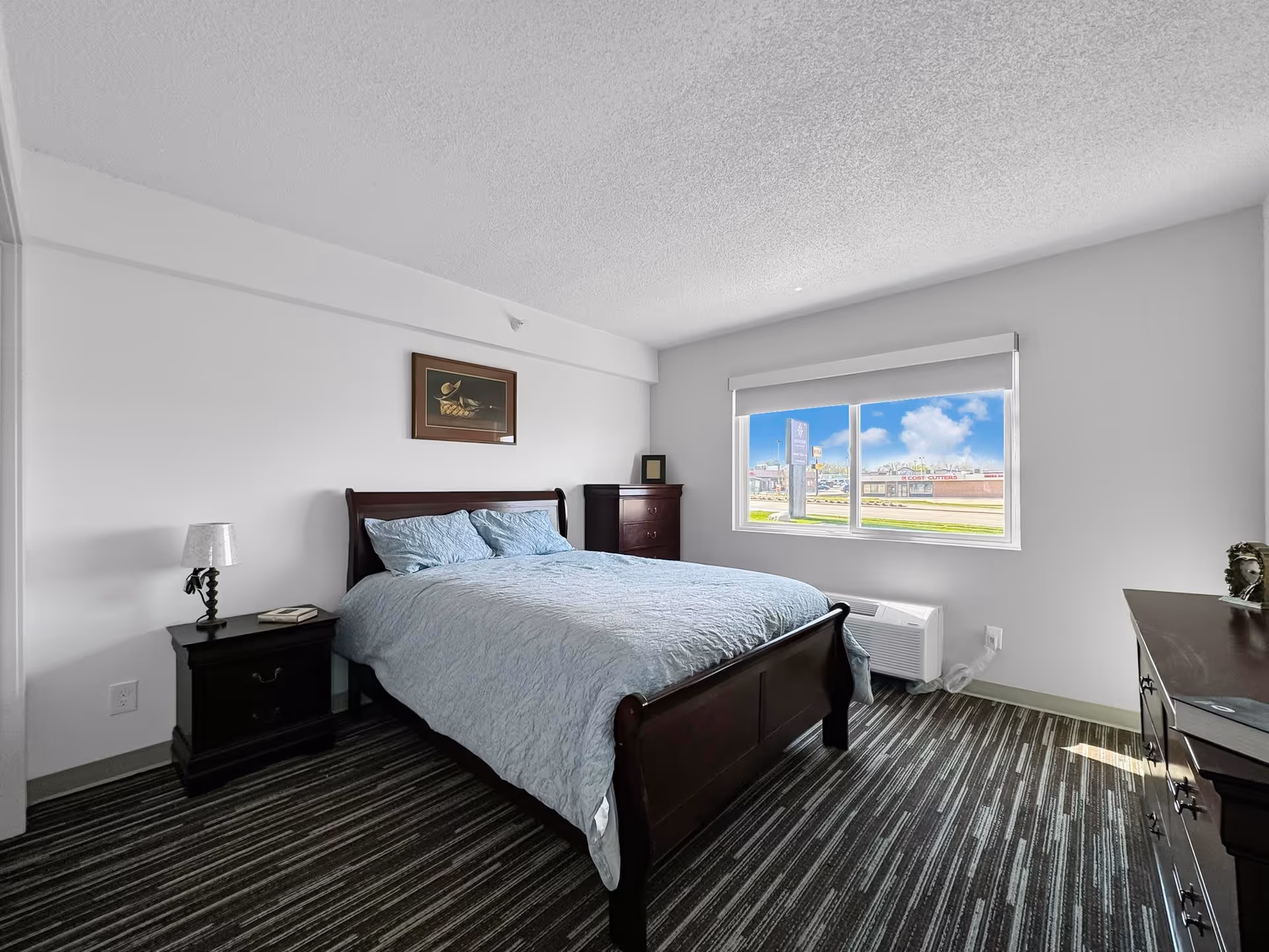 Bright bedroom with a wooden sleigh bed dressed in blue linens, nightstand, dresser, and a window overlooking the street.