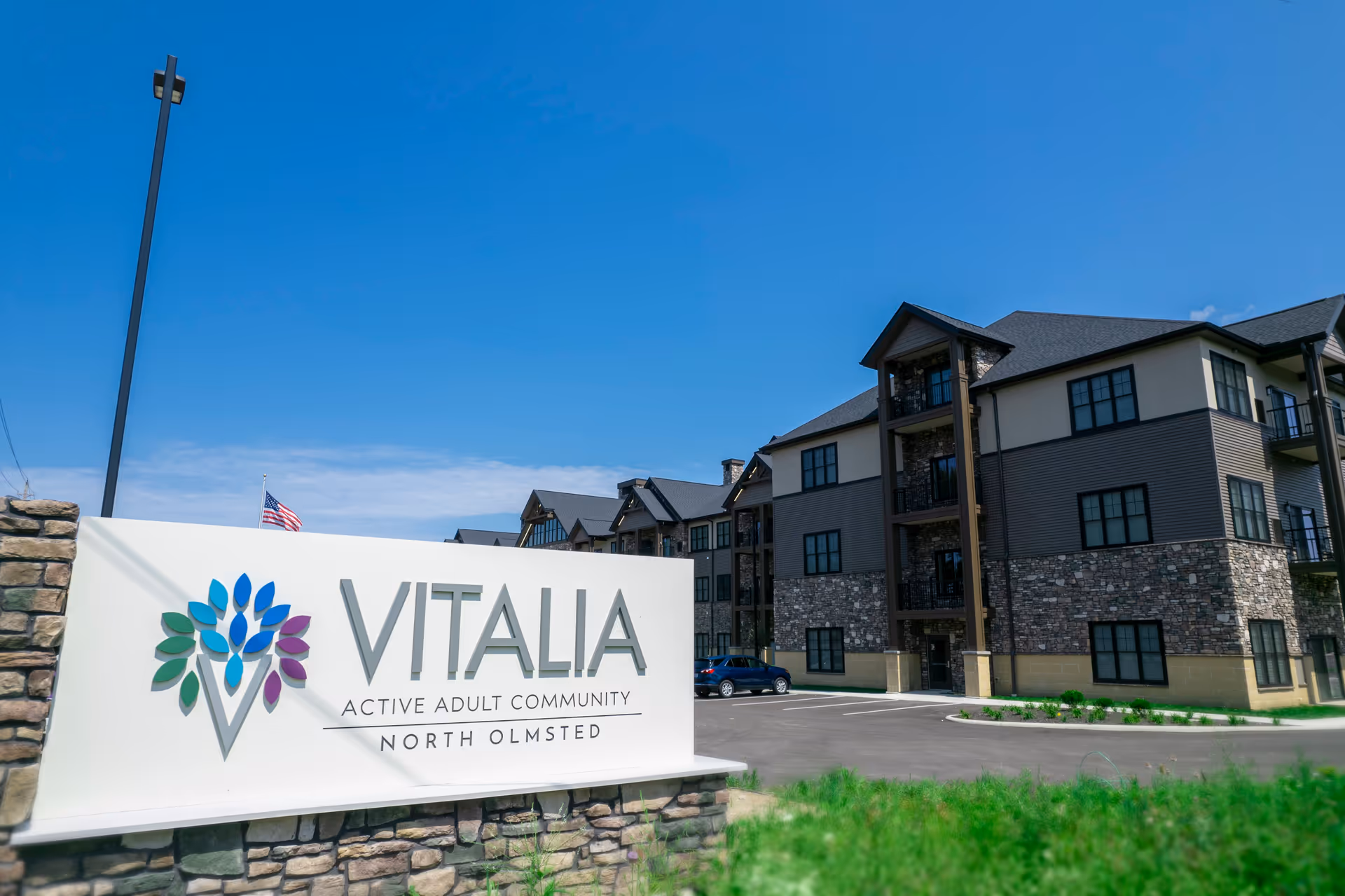 Exterior view of Vitalia Active Adult Community at North Olmsted showing a large sign with the community name and a multi-story building with stone and siding facade under a clear blue sky.