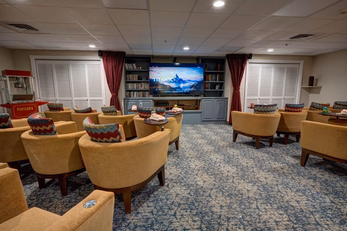 A cozy media room with multiple beige armchairs arranged in rows facing a large flat-screen TV displaying the movie Titanic. The room has patterned carpet, built-in shelves with books and DVDs, red curtains, and a popcorn machine on the left side.