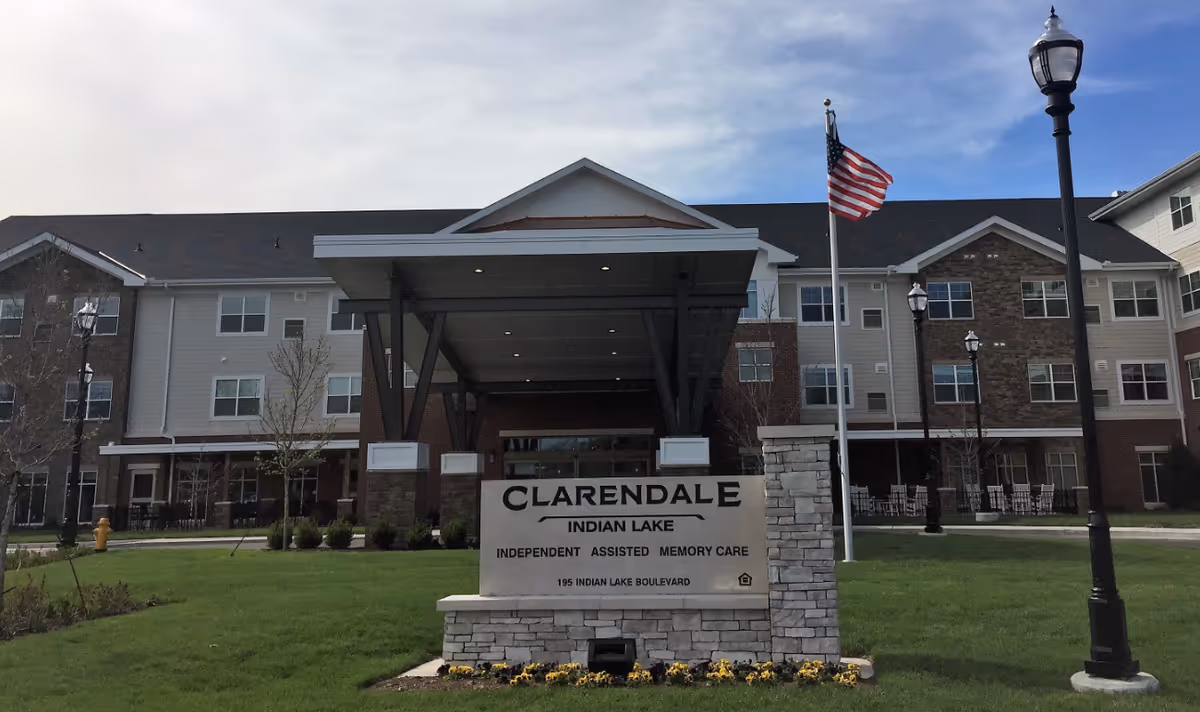 Front exterior view of Clarendale at Indian Lake senior living facility with a large covered entrance, a sign displaying the facility name and services, an American flag on a flagpole, and a well-maintained lawn with flowers and lamp posts.