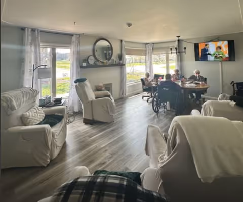 A bright living room with large windows letting in natural light. The room features several cushioned armchairs covered with white slipcovers, a round wooden table where four elderly people are seated, one in a wheelchair. A flat-screen TV is mounted on the wall showing a program. The floor is wood laminate, and there is a fireplace with a round mirror above it. Light curtains frame the windows, and the room has a calm, welcoming atmosphere.