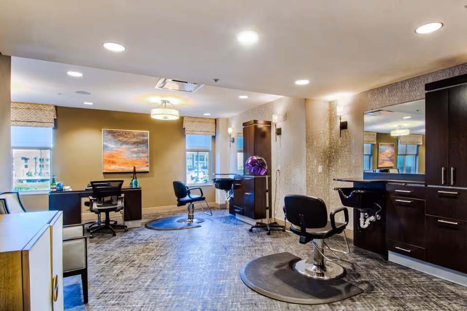 Interior view of a modern salon area in a senior living facility with two black salon chairs on mats, a large mirror, dark wood cabinetry with sinks, a desk with an office chair, and two windows with patterned valances letting in natural light. The walls have a textured wallpaper and there is a colorful abstract painting on one wall.
