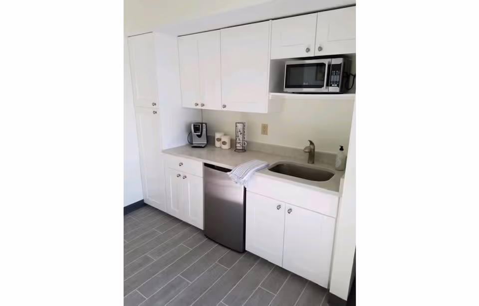 A clean and modern kitchen area with white cabinets, a stainless steel mini refrigerator, a microwave, a coffee maker, a sink with a faucet, and a countertop with a towel and some kitchen items.