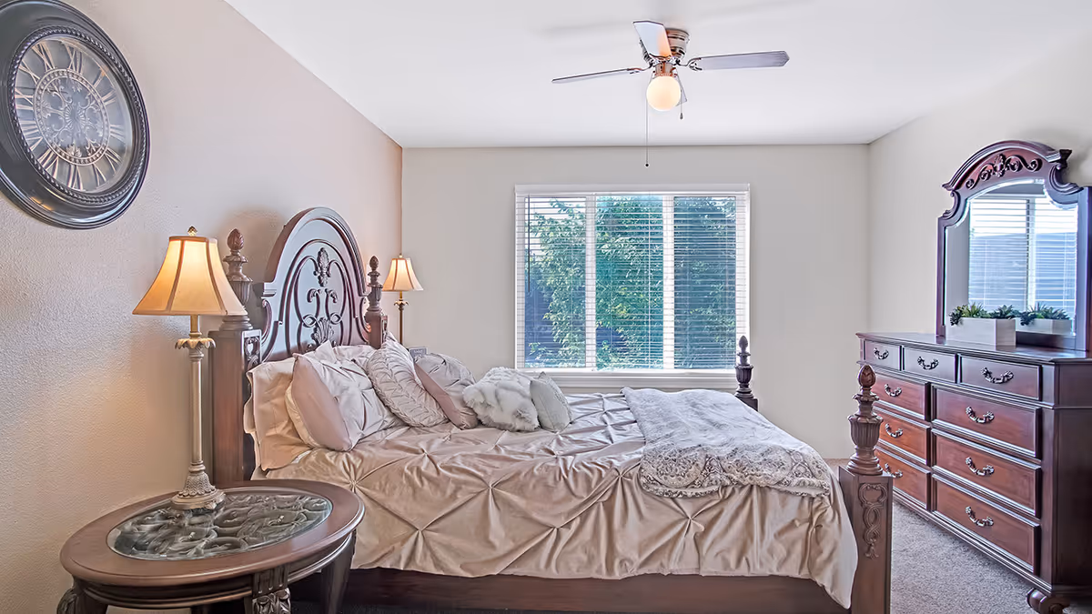A well-lit bedroom with a large wooden bed featuring an ornate headboard, beige bedding with multiple pillows, and a decorative throw. There are two bedside lamps on either side of the bed, a round side table with a glass top, a large window with white blinds showing greenery outside, and a wooden dresser with a matching ornate mirror. A ceiling fan with a light is mounted on the white ceiling.