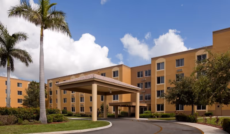 Exterior view of a multi-story senior living facility building with a covered entrance driveway, surrounded by palm trees and landscaping under a partly cloudy sky.