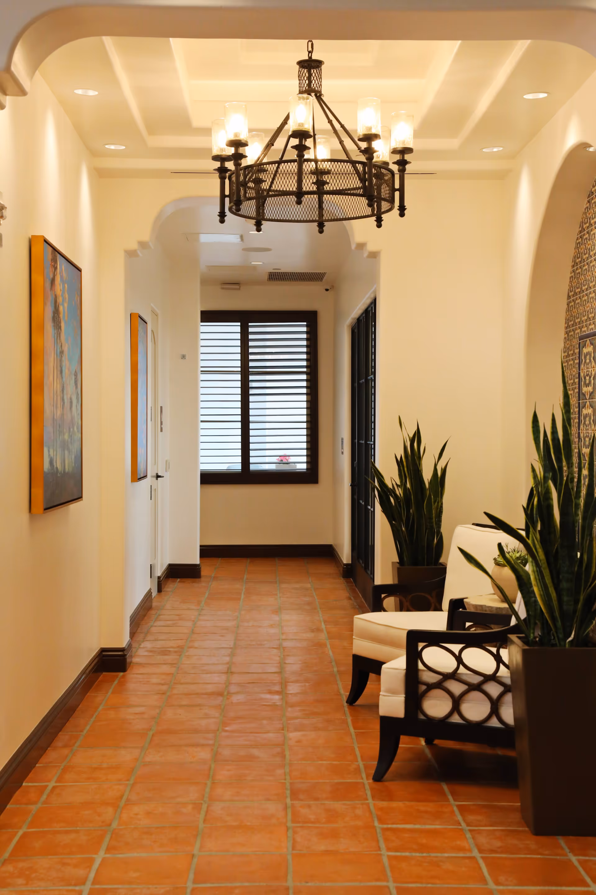 A well-lit hallway with terracotta tile flooring, cream-colored walls, and a decorative chandelier hanging from the ceiling. There are two white cushioned chairs with dark wooden frames on the right side, accompanied by tall green potted plants. A window with horizontal blinds is visible at the end of the hallway, and framed artwork hangs on the left wall.