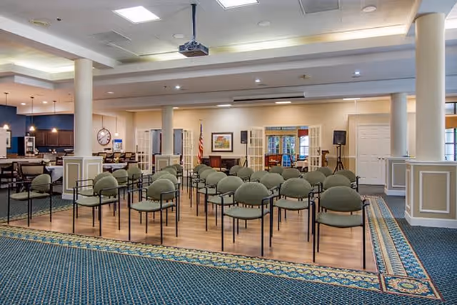 A spacious room with rows of green cushioned chairs arranged facing a small stage area with a podium and speakers. The room features a patterned blue carpet, white columns, and a ceiling-mounted projector. In the background, there is a dining area with tables and chairs, and an American flag is visible near the stage.
