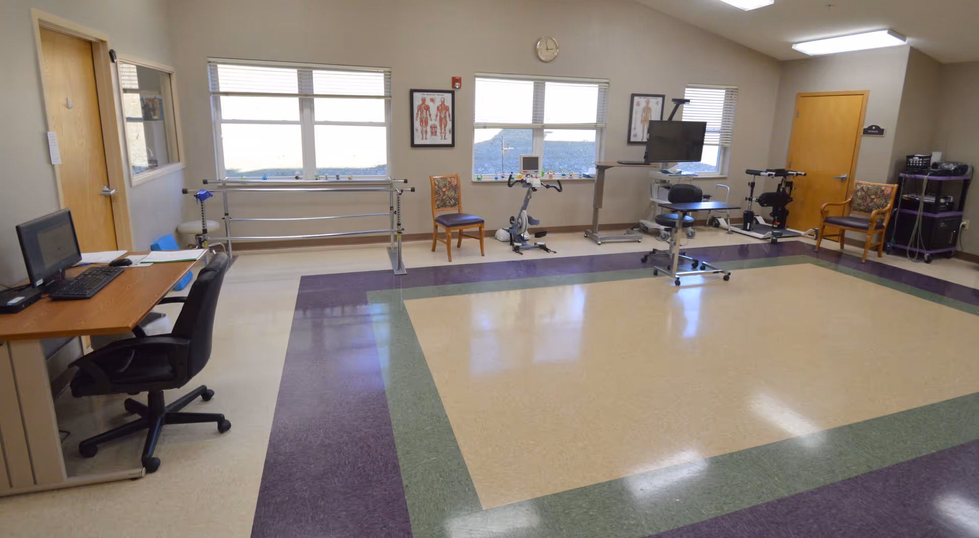 A spacious therapy or rehabilitation room with a computer desk and chair on the left, parallel bars for walking exercises along the back wall, two chairs, exercise equipment including a stationary bike, and a TV mounted on a stand. The room has large windows letting in natural light and a tiled floor with a green and purple border pattern.