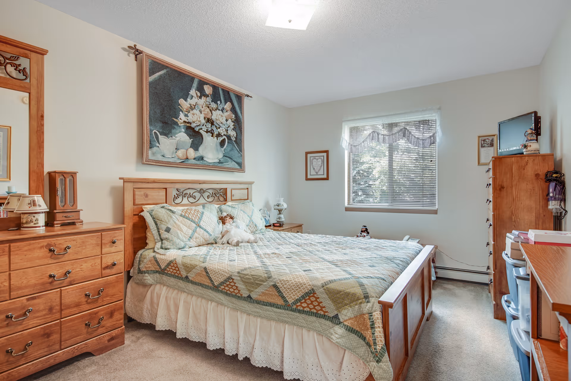 A cozy bedroom with a wooden bed frame and matching dresser and chest of drawers. The bed is made with a patchwork quilt and decorative pillows, including a doll. Above the bed hangs a tapestry featuring a floral arrangement. A window with blinds and a valance lets in natural light. The room has light-colored walls and carpeted floor.