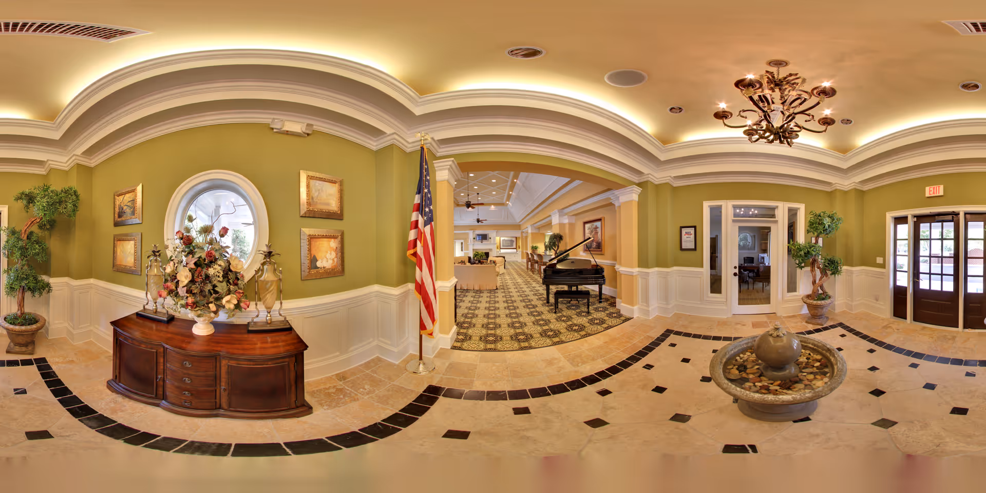A spacious and elegant senior living facility lobby with green walls, white wainscoting, and a tiled floor with black accents. The room features a wooden sideboard with a floral arrangement, framed paintings, an American flag, a grand piano, a chandelier, potted plants, and a water fountain in the center. Double glass doors lead outside.