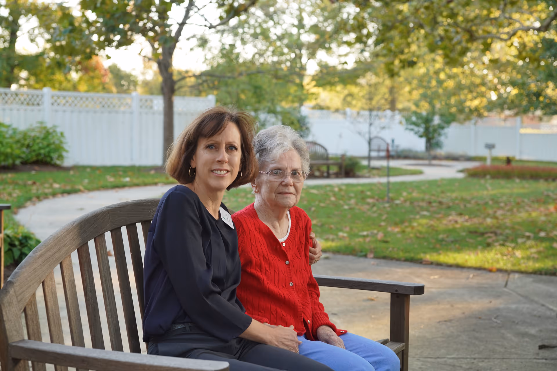 Two women sitting on a wooden bench in an outdoor garden area. One woman is elderly with gray hair, wearing glasses, a red sweater, and blue pants. The other woman has brown hair and is wearing a dark long-sleeve top. They are surrounded by green grass, trees, and a white fence in the background.