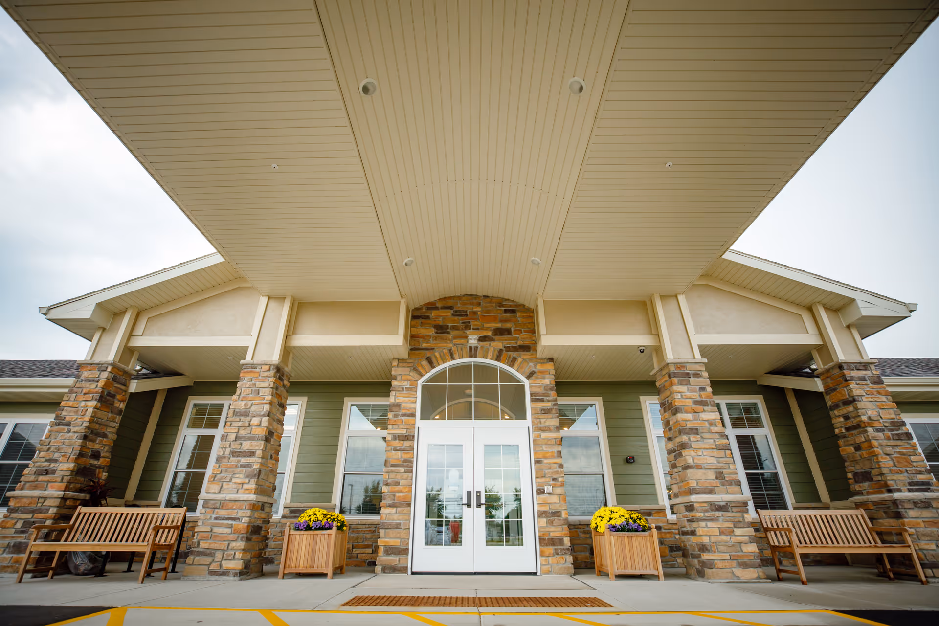 Front entrance of The Courtyard at Oshkosh Assisted Living and Memory Care facility featuring a covered porch with stone pillars, double glass doors, two wooden benches, and flower planters on either side of the entrance.