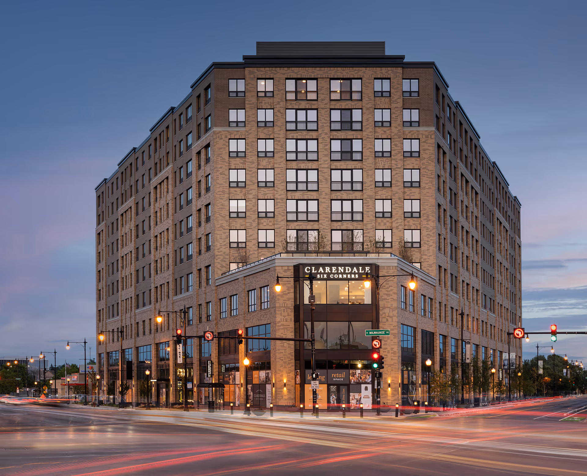 Exterior view of a large multi-story brick building named Clarendale Six Corners at dusk, with street lights and traffic light trails visible at the intersection in front.