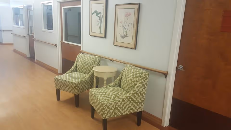 Hallway seating area with two green patterned chairs and a small round table against a wall with handrails and framed botanical prints.