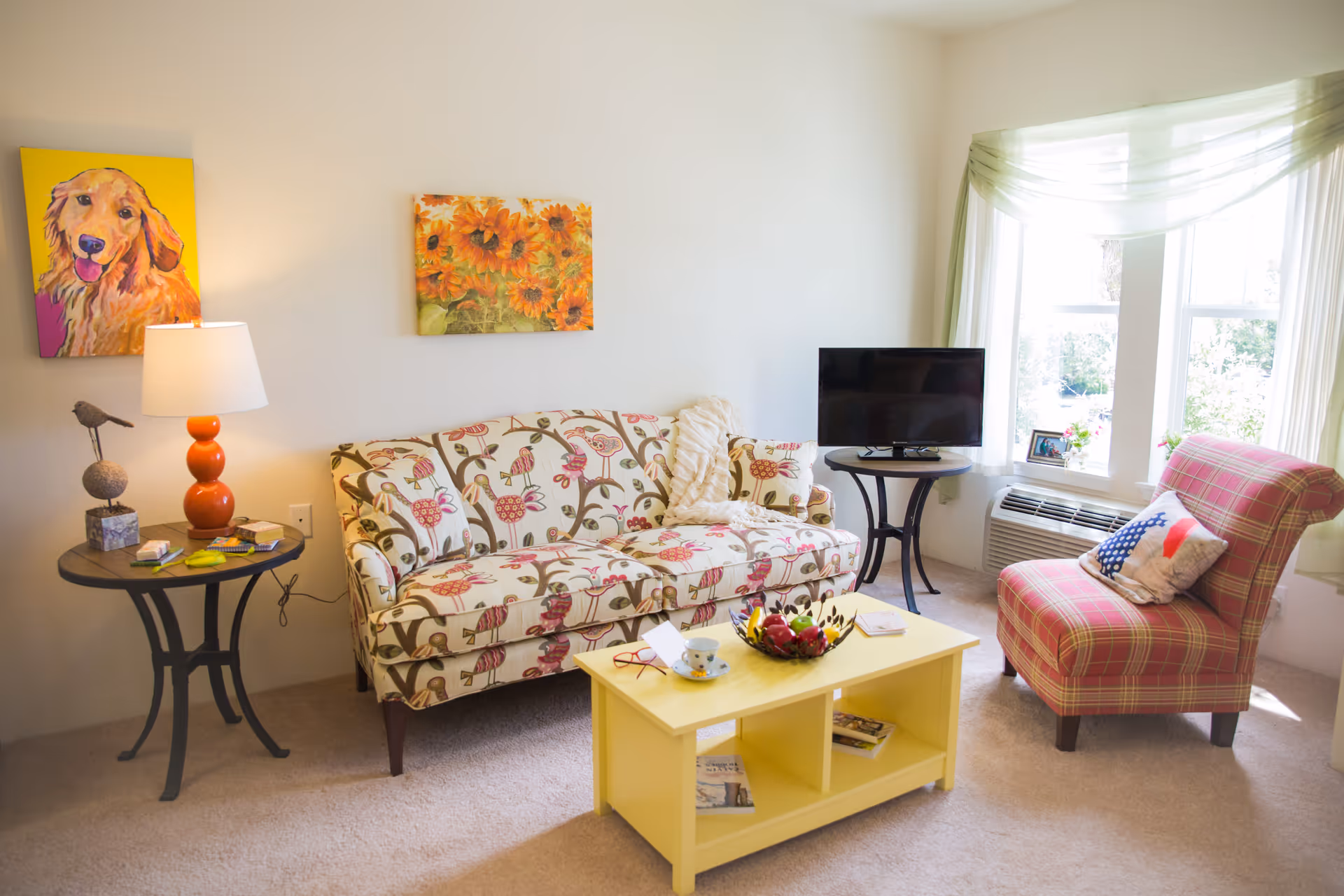 A cozy living room with a floral patterned sofa, a red plaid armchair with a decorative pillow, a small yellow coffee table with a teacup and a decorative bowl, a round side table with an orange lamp and small decor items, a flat-screen TV on a small black table, and two colorful paintings on the wall. The room has a large window with light green sheer curtains letting in natural light.