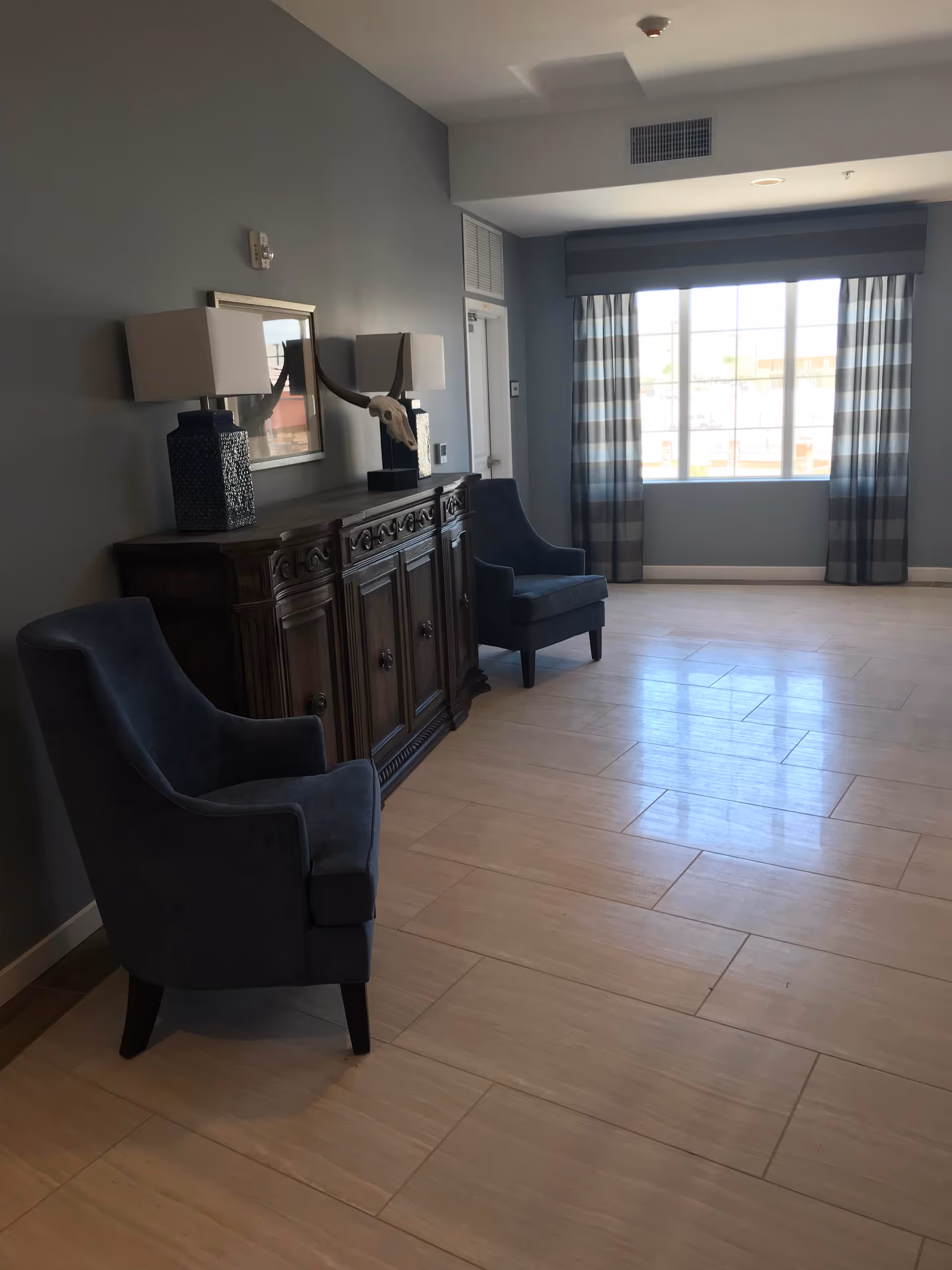 A room with two dark blue upholstered armchairs placed on either side of a wooden sideboard with decorative carvings. On top of the sideboard are two lamps with rectangular shades and a decorative skull with horns. A large window with striped curtains allows natural light to fill the room, which has light-colored tiled flooring and gray walls.