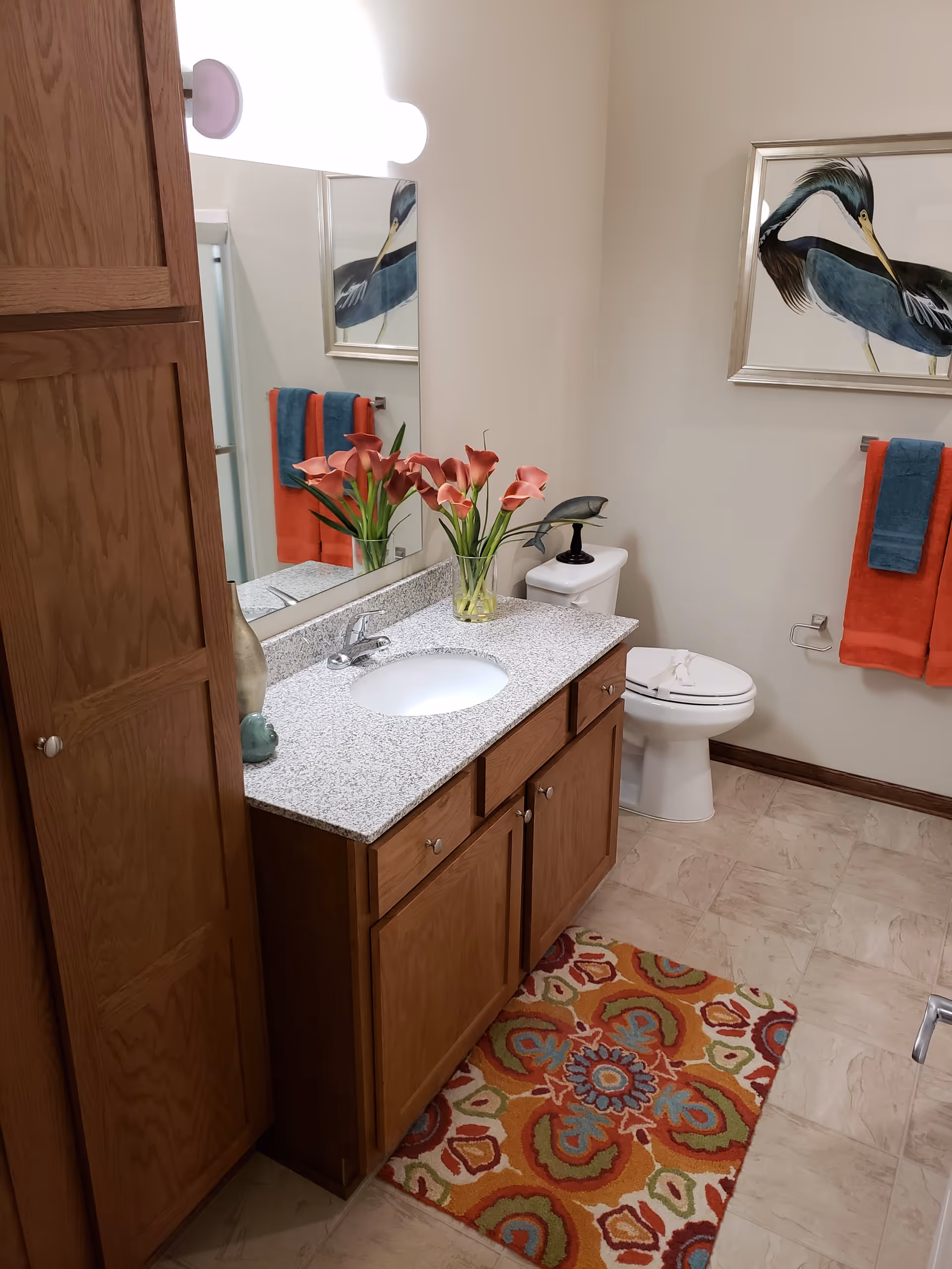 A bathroom with a wooden cabinet and granite countertop with a sink. There is a vase with pink flowers on the countertop. A toilet is visible next to the cabinet, with a decorative bird sculpture on top of the toilet tank. Two colorful towels, one orange and one blue, hang on a towel rack on the wall. Above the towels is a framed picture of a bird. The floor has a colorful patterned rug.