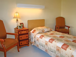 A small bedroom with a single bed covered in a patterned quilt. Next to the bed is a wooden nightstand with three drawers, a table lamp, and framed photos. Two wooden armchairs with orange cushions are positioned on either side of the nightstand. The walls are light-colored and the floor is carpeted.