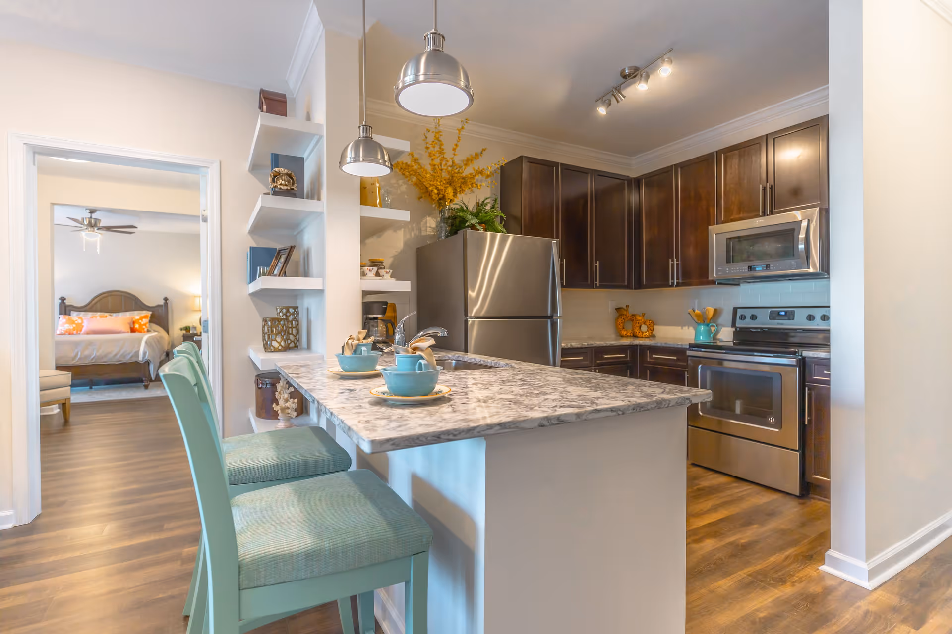 Modern kitchen with dark wooden cabinets, stainless steel refrigerator, oven, and microwave. A marble countertop island with two teal cushioned chairs and teal dishware is in the foreground. Open shelves with decorative items are on the left wall. A bedroom with a bed and ceiling fan is visible through an open doorway.