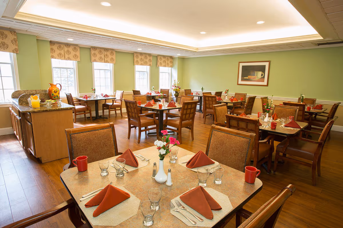 Bright dining room with multiple set tables, wooden chairs, and place settings in a senior living facility.