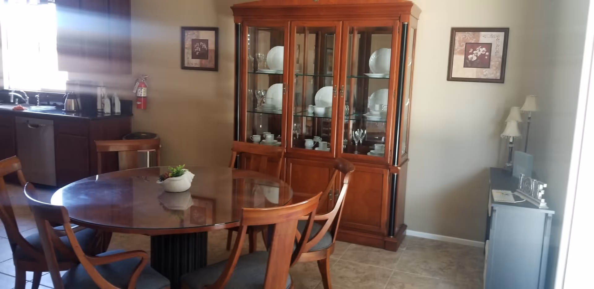A dining area with a round wooden table surrounded by four wooden chairs with cushions. Behind the table is a wooden china cabinet displaying plates, bowls, and glassware. To the left, part of a kitchen with a sink, dishwasher, and countertop appliances is visible. On the right side, there is a small cabinet with two table lamps and framed artwork on the walls.