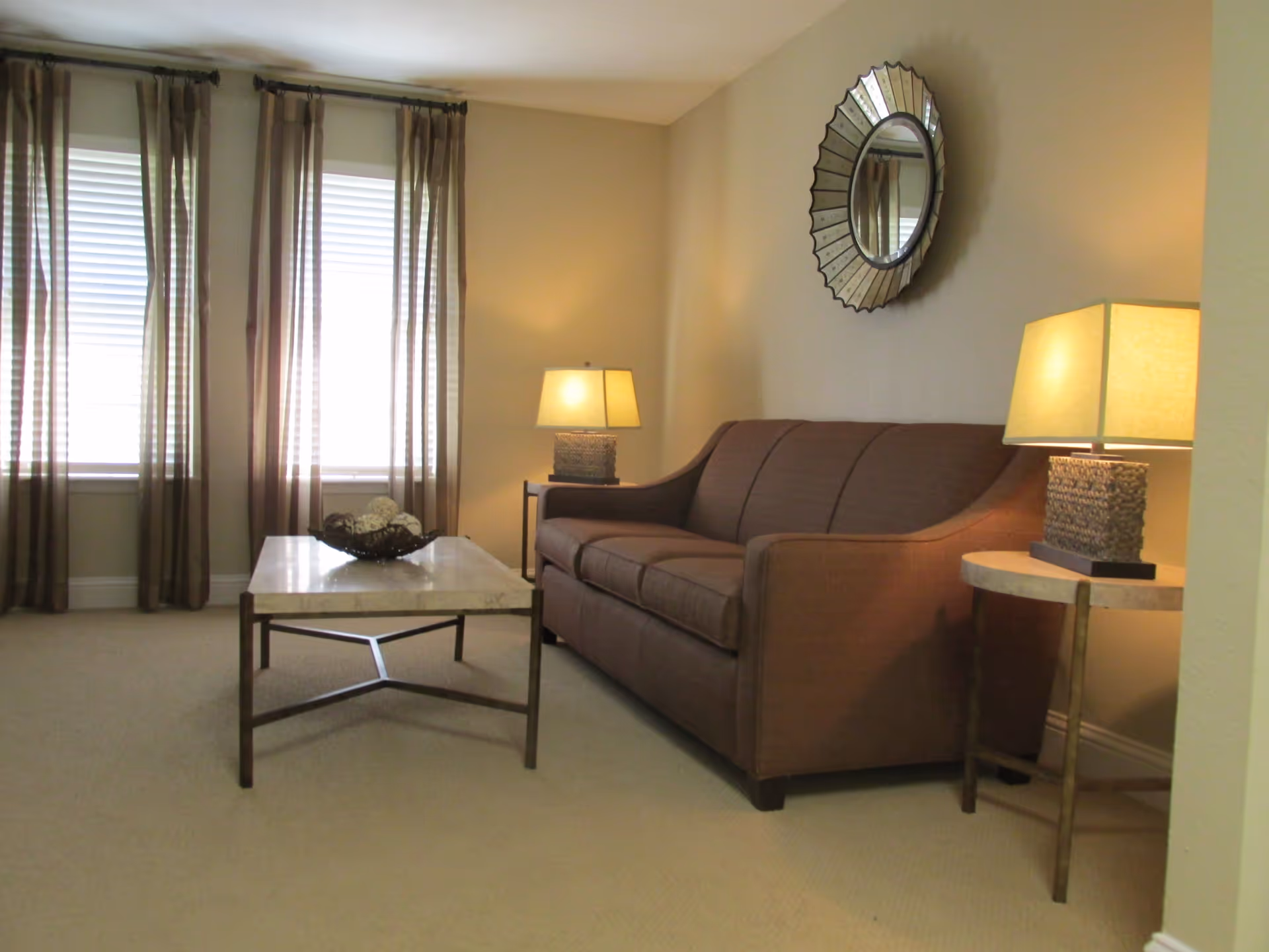 A cozy living room with a brown sofa, two side tables each holding a lamp, a rectangular coffee table with decorative items, sheer brown curtains covering two windows, and a round decorative mirror on the wall.