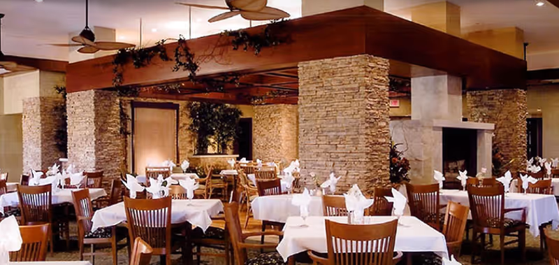 A spacious dining room with multiple tables covered in white tablecloths, each set with folded napkins and glassware. The room features stone pillars, wooden ceiling beams with greenery, ceiling fans, and a fireplace in the background.