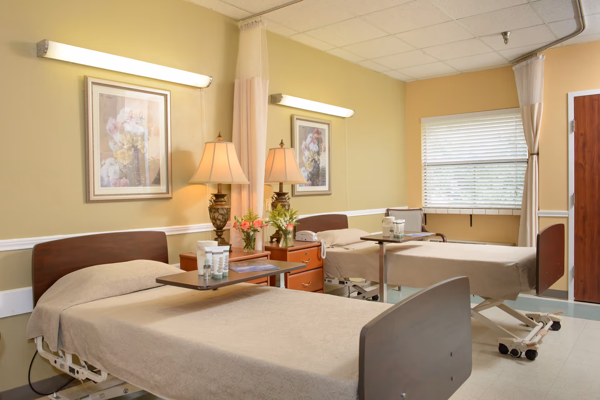 A room with two hospital-style beds covered with beige blankets, each with an overbed table holding personal care items. Between the beds is a wooden nightstand with two decorative lamps and flower arrangements. The walls are painted light yellow with framed floral artwork, and a window with blinds is visible in the background.