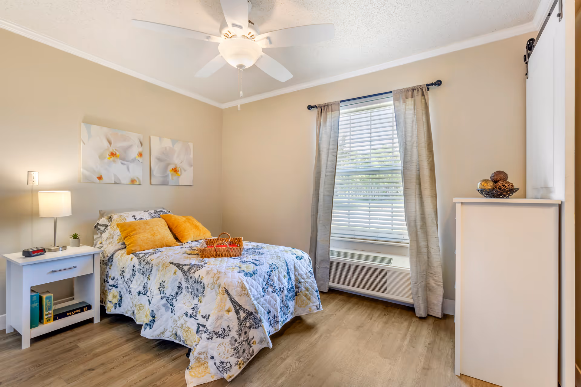 A bright, tidy bedroom with a floral-patterned bed topped with yellow pillows and a wicker tray, a nightstand with lamp, a window with blinds and curtains, and a ceiling fan.