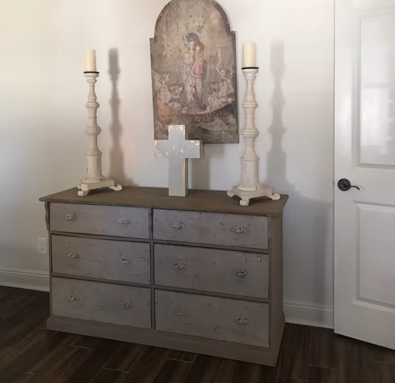 A distressed gray six-drawer dresser topped with tall candlesticks, a wooden cross, and a religious wall plaque next to a white door.