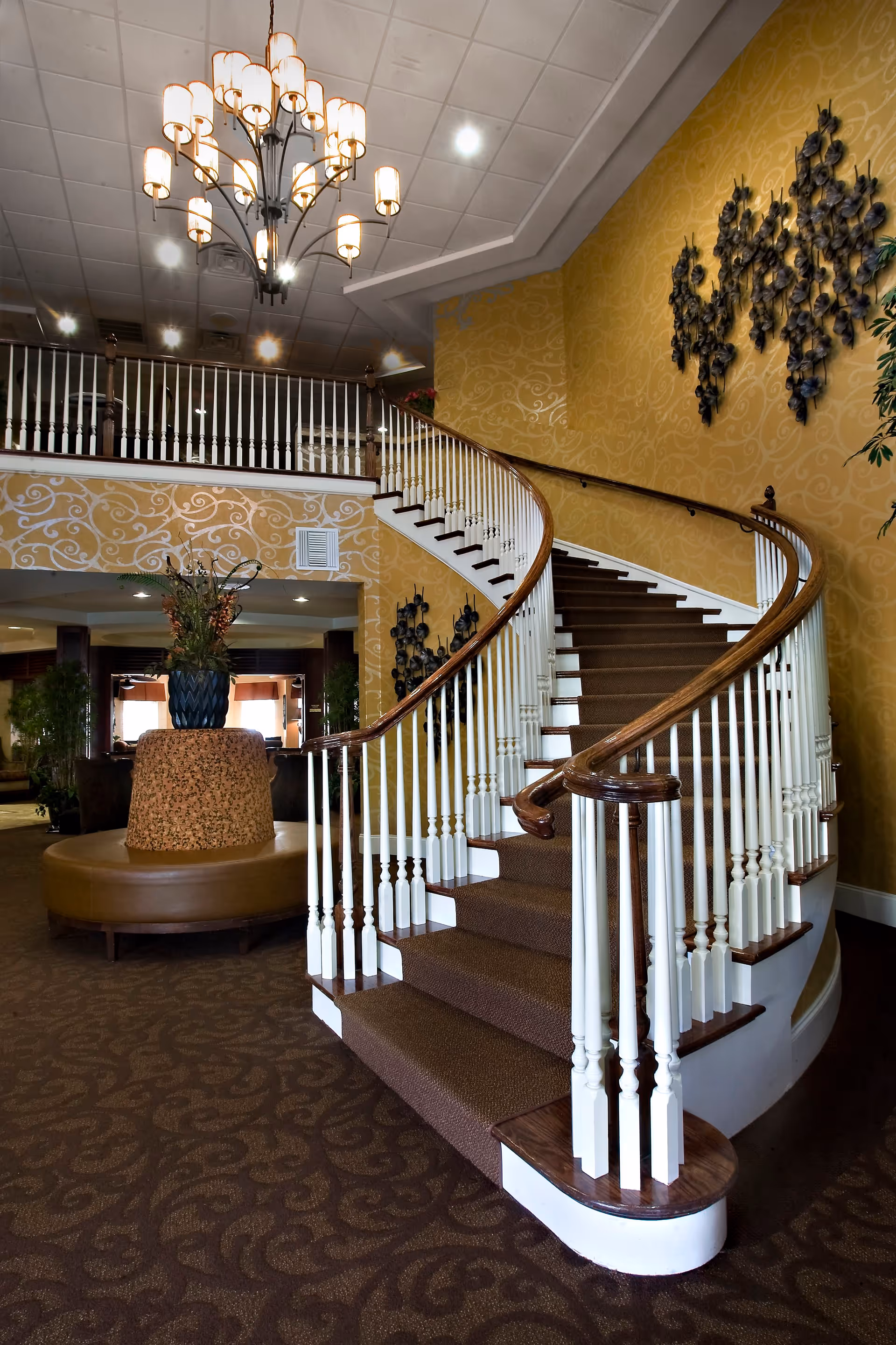 Interior view of a senior living facility featuring a grand curved staircase with white balusters and wooden handrails. The walls are decorated with a yellow patterned wallpaper and metal wall art. A large chandelier hangs from the ceiling, and there is a round cushioned bench with a decorative vase and plants in the center of the room. The carpet has a dark, intricate pattern.