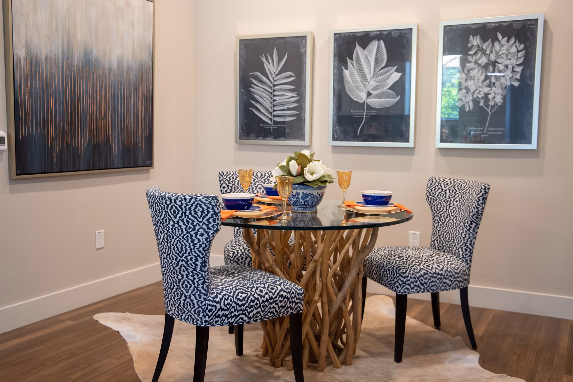 A dining area with a round glass table supported by a base made of intertwined wooden branches. Four upholstered chairs with a blue and white geometric pattern surround the table. The table is set with blue bowls, orange napkins, and amber-colored glasses. A floral centerpiece in a blue and white vase is on the table. The walls have three framed botanical prints and one abstract painting. The floor is wooden with a light-colored rug underneath the table.