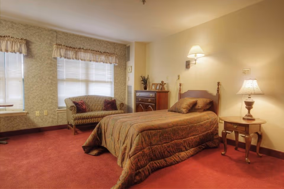 A cozy bedroom in Stratford Commons Assisted Living featuring a single bed with a patterned bedspread and pillows, a wooden nightstand with a lamp, a wooden dresser with decorative items, a small upholstered loveseat with two cushions, and two windows with blinds and valances. The room has warm lighting and a red carpeted floor.