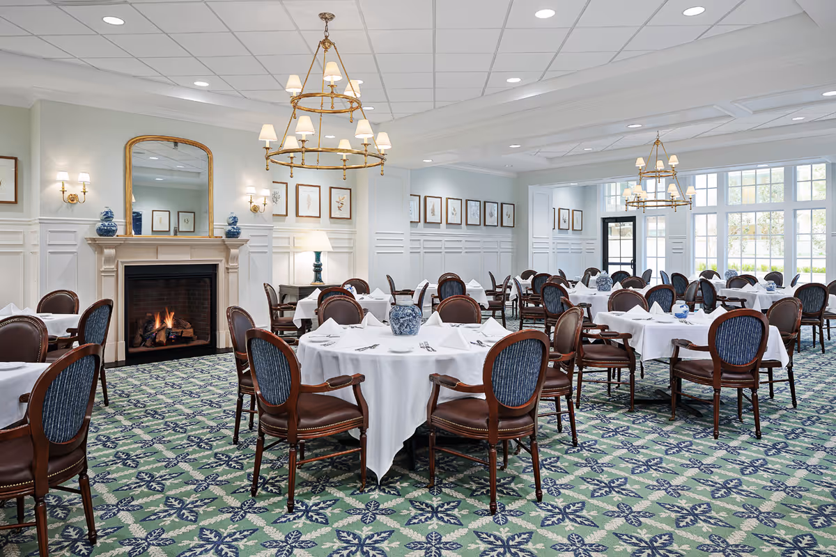 A spacious dining room with round tables covered in white tablecloths, each set with napkins, silverware, and blue and white decorative vases. The room features elegant wooden chairs with blue upholstery, a fireplace with a lit fire, a large mirror above the fireplace, framed artwork on the walls, and two gold chandeliers hanging from a white ceiling. Large windows allow natural light to fill the room.