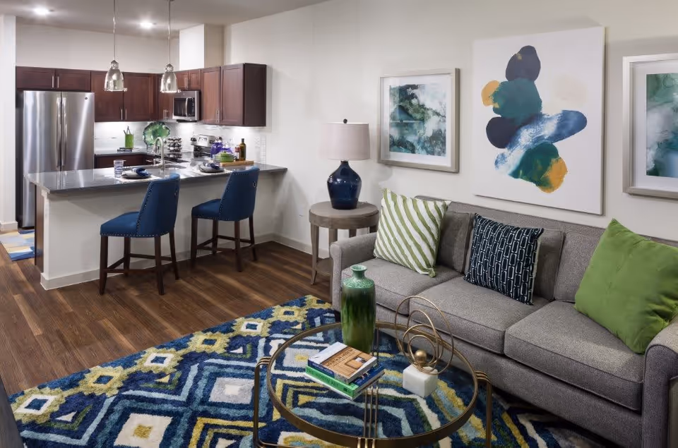 A modern open-concept living room and kitchen area with wood flooring. The living room features a gray sofa with green and patterned pillows, a round glass coffee table with decorative items and books, and colorful abstract artwork on the walls. The kitchen has dark wood cabinets, stainless steel appliances, a granite countertop island with two blue upholstered bar stools, and pendant lighting above the island.