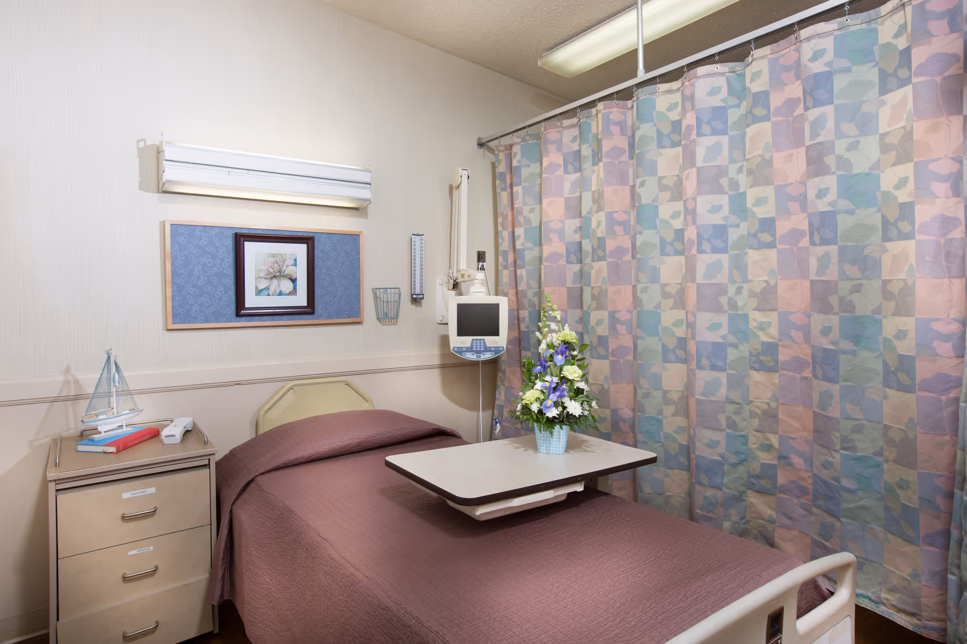 A rehabilitation patient room with a single bed, overbed table holding a vase of flowers, medical monitor, bedside drawers and a patterned privacy curtain.