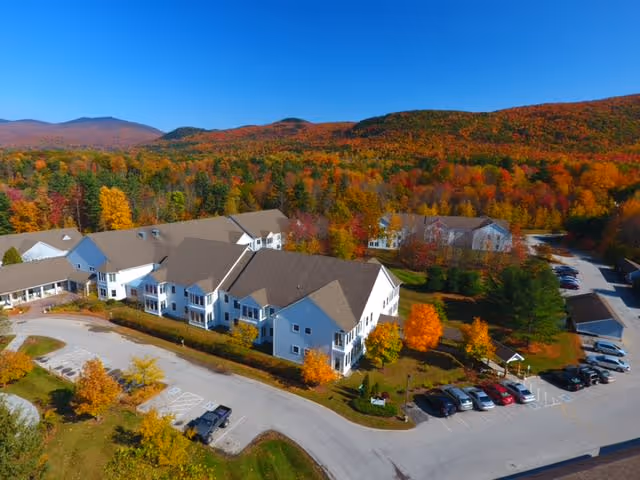 Aerial view of a senior living facility named Gables at East Mountain surrounded by colorful autumn trees and hills in the background. The building is white with a brown roof, and there is a parking lot with several cars parked. The landscape features vibrant fall foliage with shades of orange, yellow, and red.