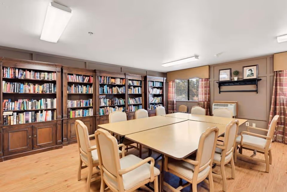 A well-lit room with a large wooden bookshelf filled with books along one wall. In the center, there are four tables pushed together forming a square, surrounded by cushioned chairs with wooden frames. The room has wooden flooring, beige walls with a window covered by plaid curtains, and a small shelf with framed pictures and a plant above an air conditioning unit.