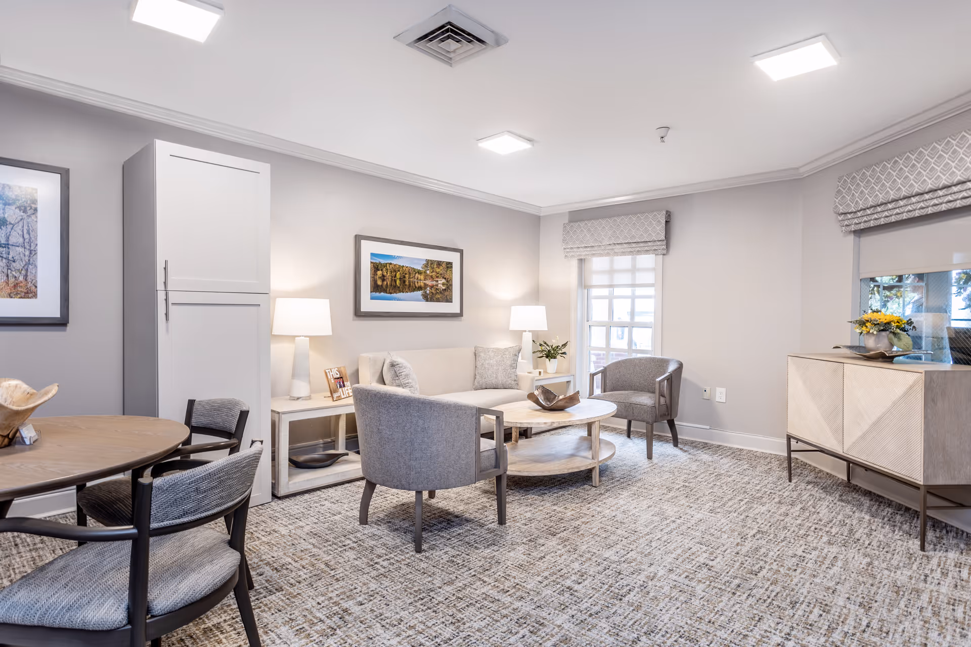 A bright and cozy living room area with light gray walls and carpeted floor. The room features a round wooden coffee table surrounded by a gray loveseat and two gray armchairs. There are two table lamps on side tables, framed landscape artwork on the walls, a wooden cabinet with decorative items, and a round dining table with chairs in the foreground. Windows with patterned Roman shades allow natural light into the space.
