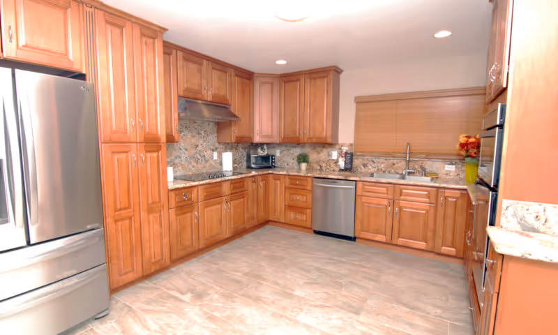A spacious kitchen with wooden cabinets, stainless steel refrigerator, dishwasher, and oven. The countertops and backsplash are made of a matching stone material. There is a window with closed blinds above the sink and recessed lighting in the ceiling.