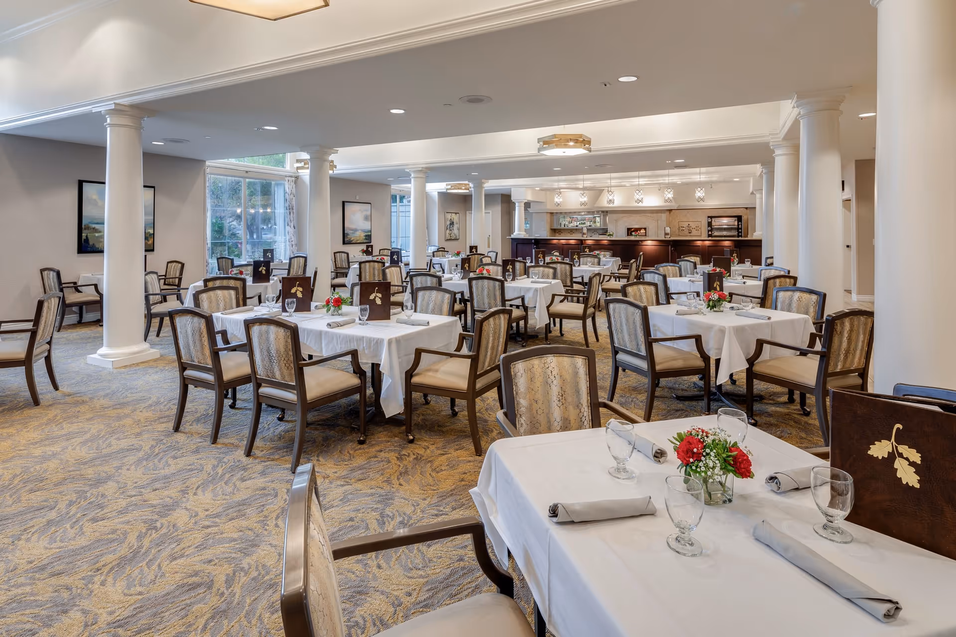 A spacious dining room with multiple tables covered in white tablecloths, each set with glassware, napkins, and small floral centerpieces. The room features beige patterned carpet, white columns, and large windows allowing natural light to enter. The back of the room has a serving area with warm lighting and decorative elements.