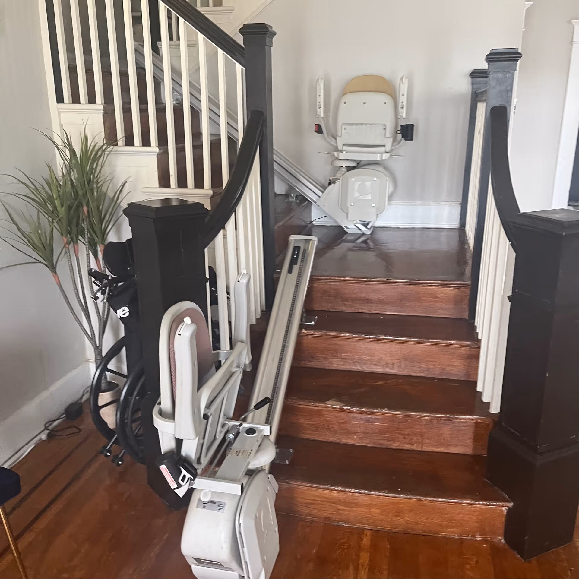 Wooden staircase with a stair lift installed on the right side. The stair lift has a beige seat and armrests. A wheelchair is positioned near the base of the stairs next to a potted plant with long green leaves. The staircase has white spindles and dark wooden handrails and newel posts.
