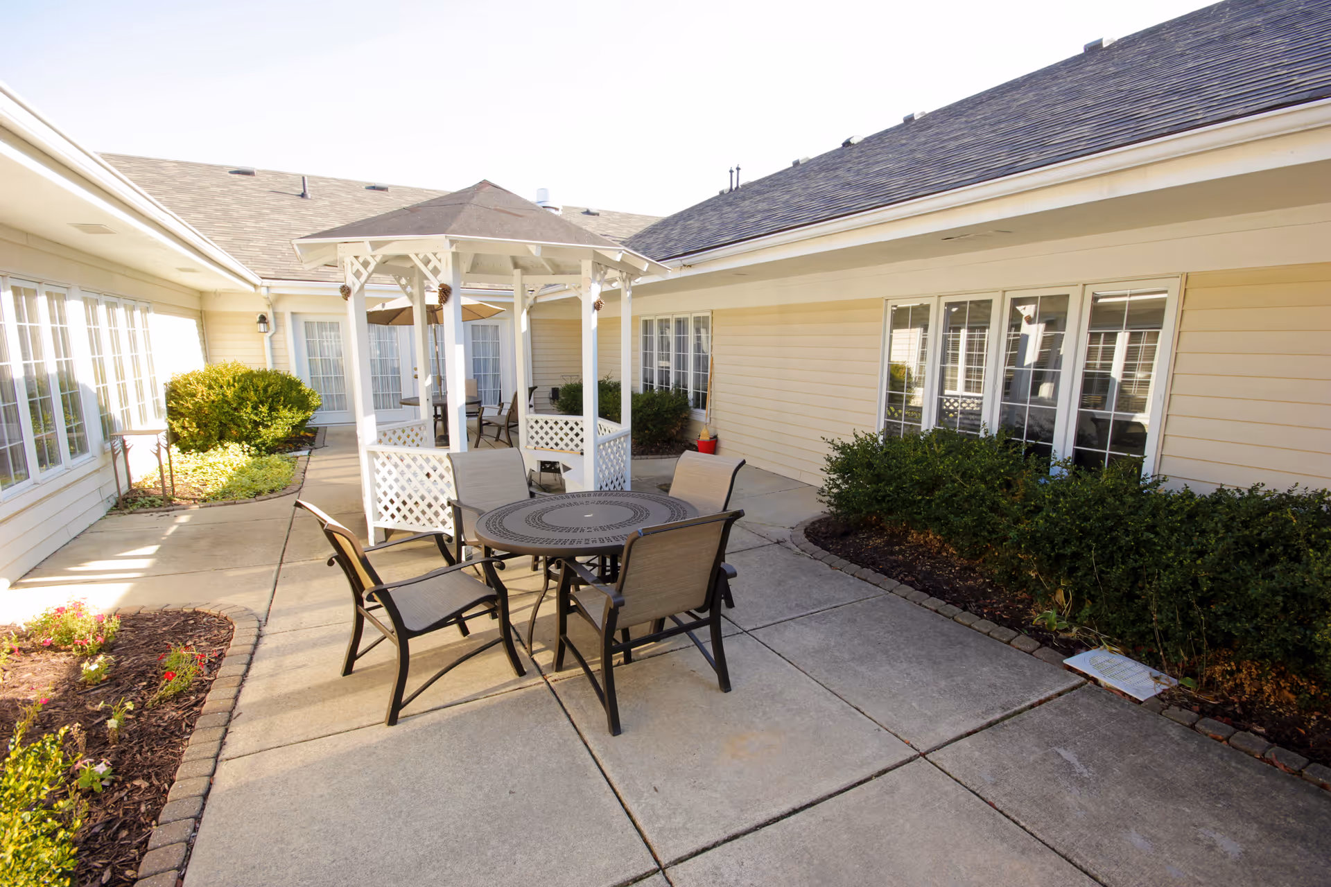 Outdoor courtyard area with a round metal table and four chairs on a concrete patio. There is a white gazebo with lattice sides and additional seating inside. The courtyard is surrounded by beige building walls with multiple windows and some green bushes and plants along the edges.