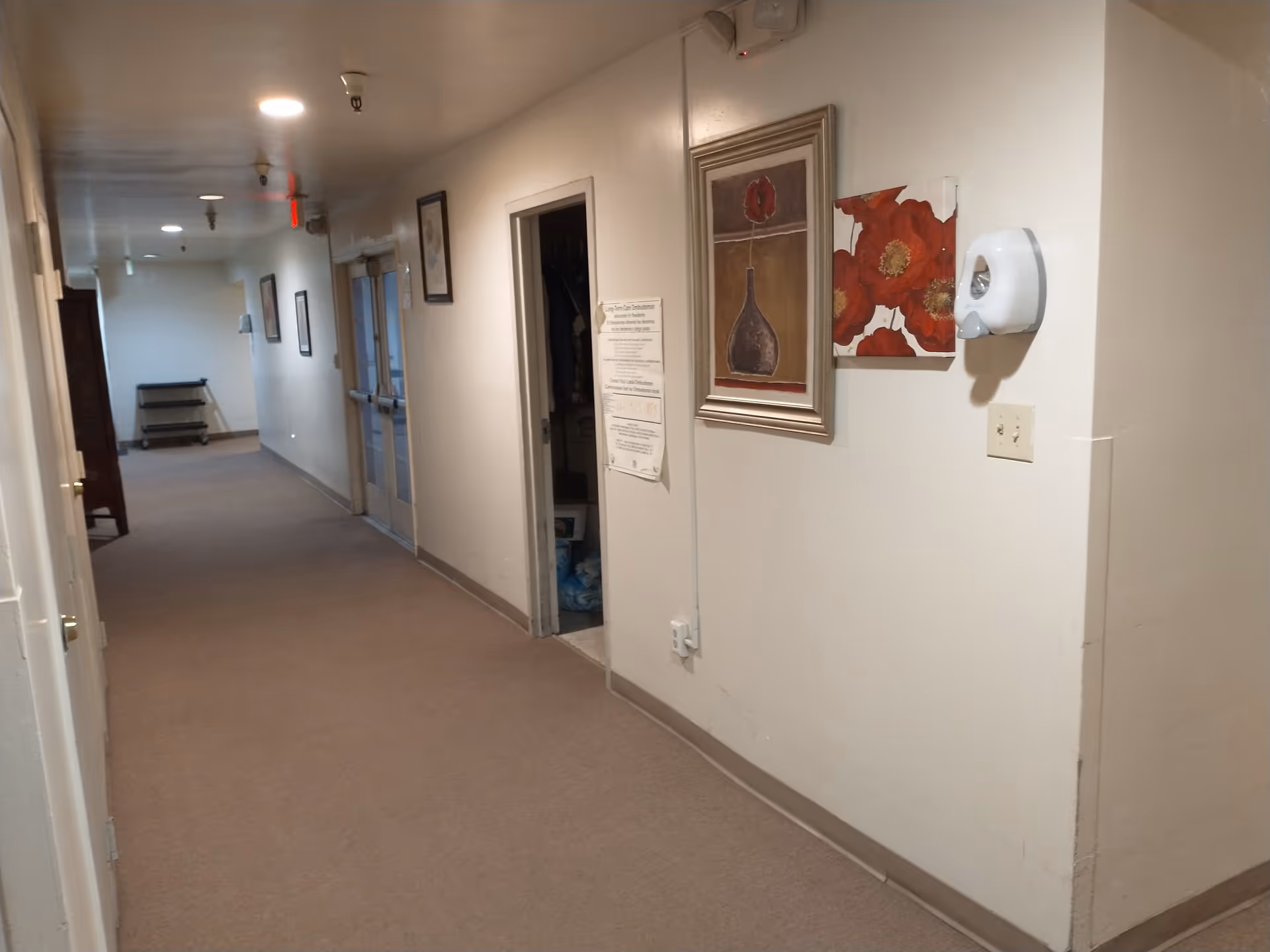 Carpeted interior hallway in an assisted living facility with framed artwork on the wall and an open doorway.