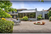 Outdoor courtyard area of a retirement and assisted living facility with patio seating, greenery, bushes, and a building with large windows under a partly cloudy sky.
