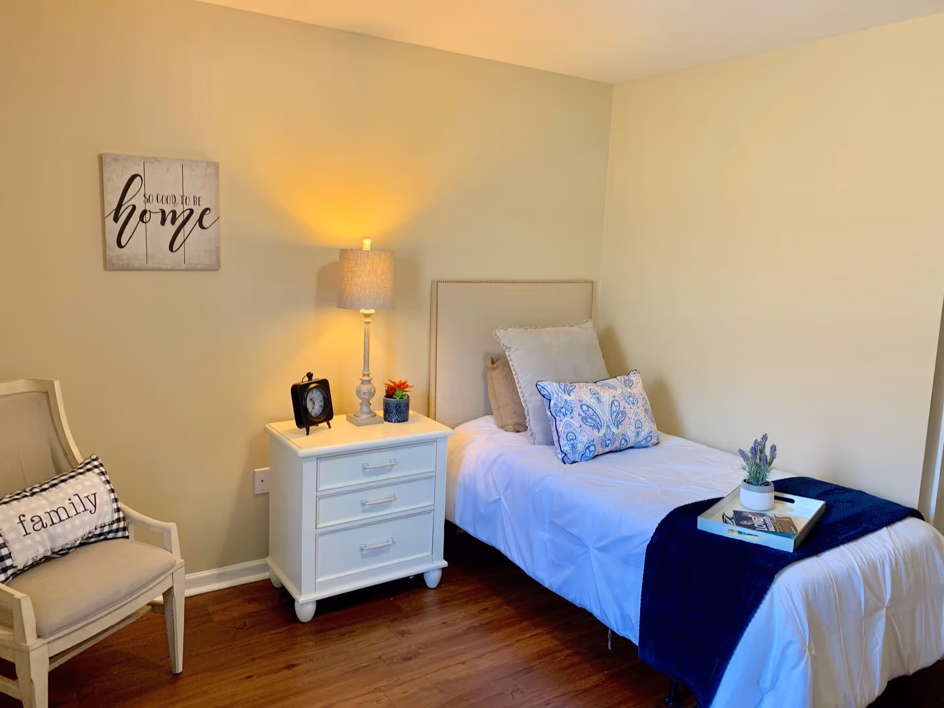 A cozy bedroom with a single bed dressed in white bedding and multiple pillows, including a blue patterned one. A navy blue throw blanket is folded at the foot of the bed with a tray holding a small potted plant and magazines. Next to the bed is a white nightstand with three drawers, a table lamp, a small clock, and a small plant. To the left is a beige armchair with a black and white checkered pillow that says 'family'. On the wall above the nightstand is a decorative sign that reads 'So good to be home'. The room has wooden flooring and light-colored walls.