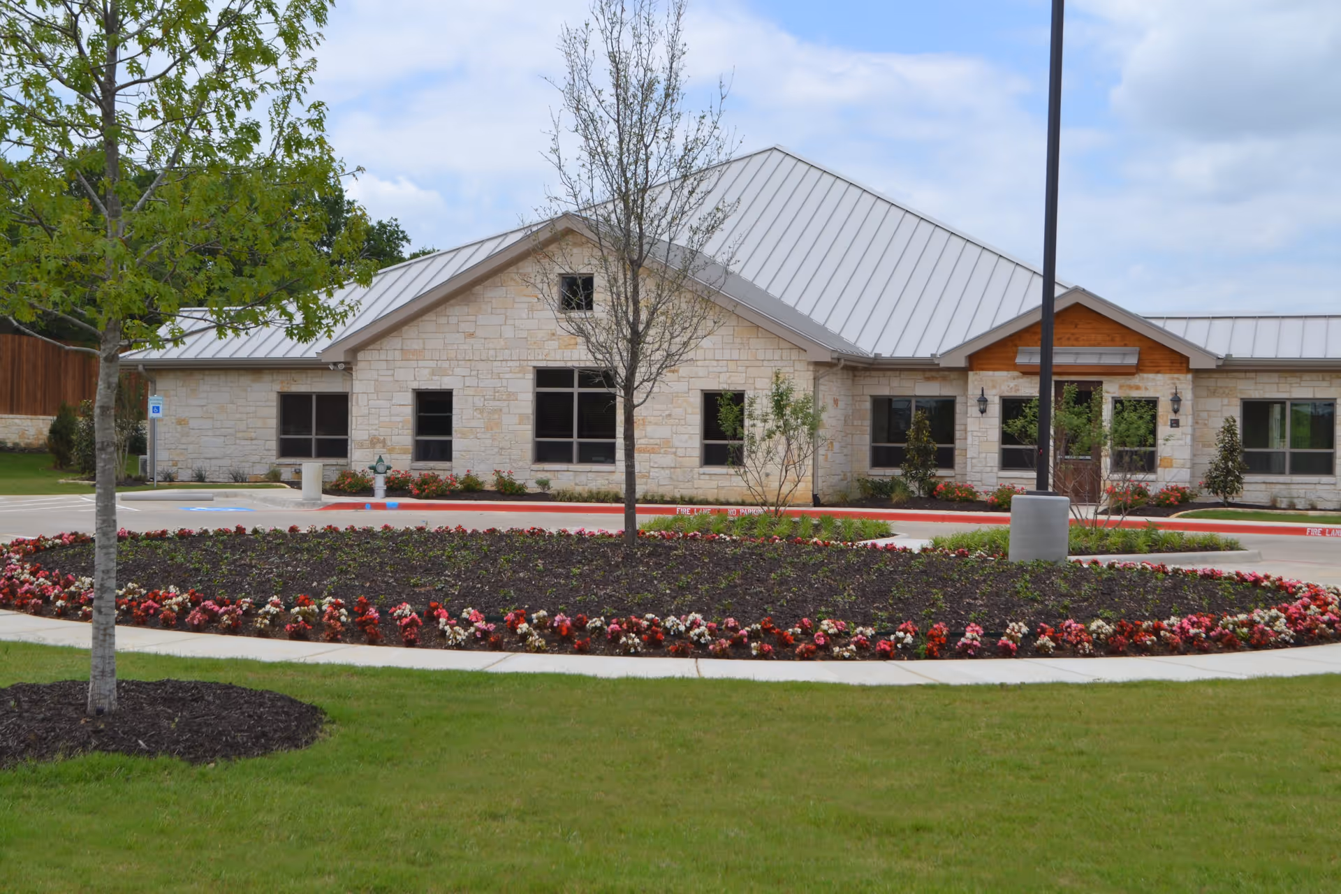 Exterior view of a single-story assisted living facility building with light-colored stone walls and a metal roof. In front of the building is a circular flower bed with red and pink flowers, surrounded by a sidewalk and green grass. There are a few young trees and a lamppost near the flower bed. The sky is partly cloudy.