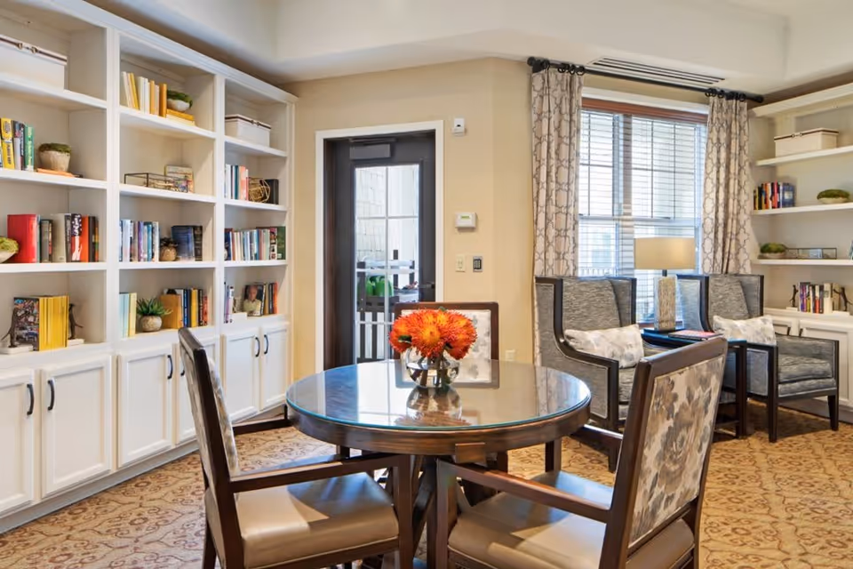 A cozy interior room featuring a round wooden table with a glass top and four cushioned chairs, two with floral upholstery. The room has built-in white bookshelves filled with books and decorative plants. There are two armchairs with patterned cushions near a window with patterned curtains, and a table lamp on a small side table between them. A door with a glass panel is visible in the background.