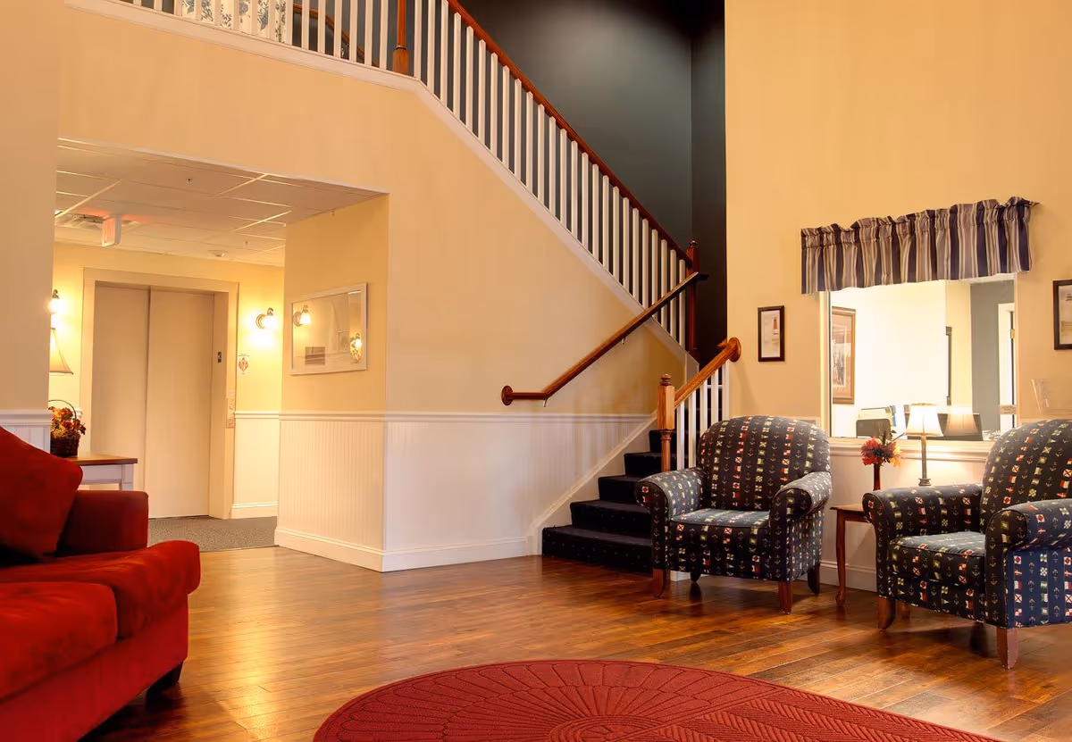 A cozy interior common area with wooden flooring, a red sofa on the left, two patterned armchairs on the right, a small table with a lamp and flowers between the chairs, and a staircase with white railings leading upstairs. There is a window with a striped valance above the chairs and soft lighting on the walls.