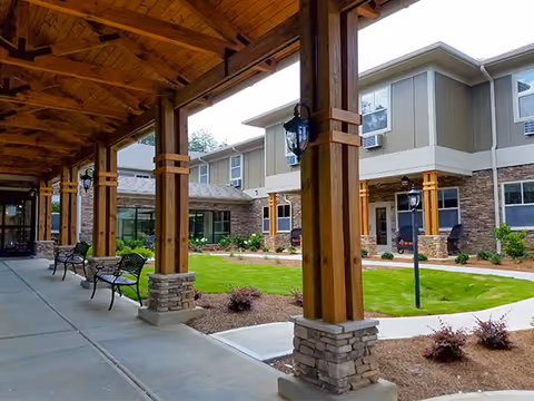 Covered walkway with wooden beams and stone bases overlooking a landscaped courtyard with green grass, shrubs, and a two-story senior living building in the background.